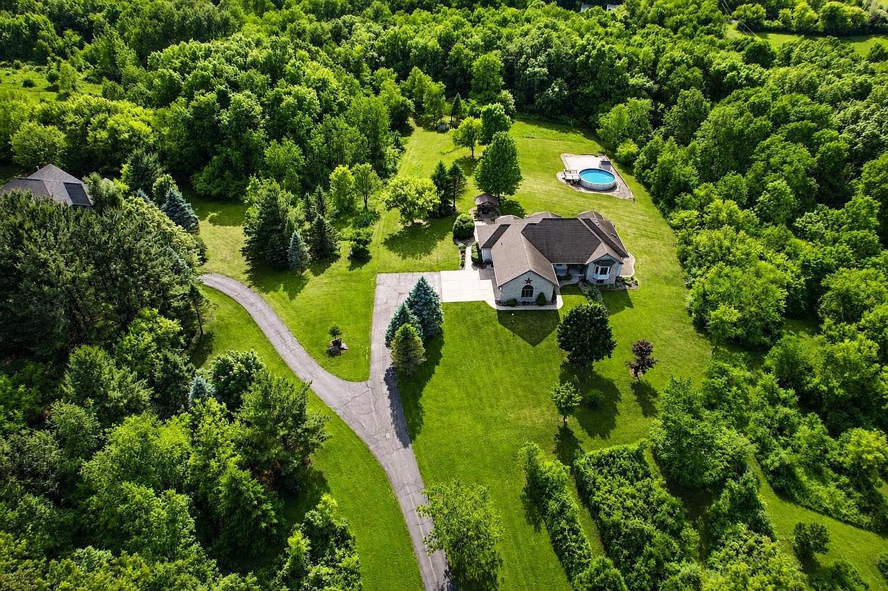 High aerial of 1825 Bringold Drive in summer: the custom home set right-of-center on its 5.79 acres, the round above-ground pool tucked into the lawn at the upper right, and a private paved driveway curving in from the lower left through a wall of mature trees