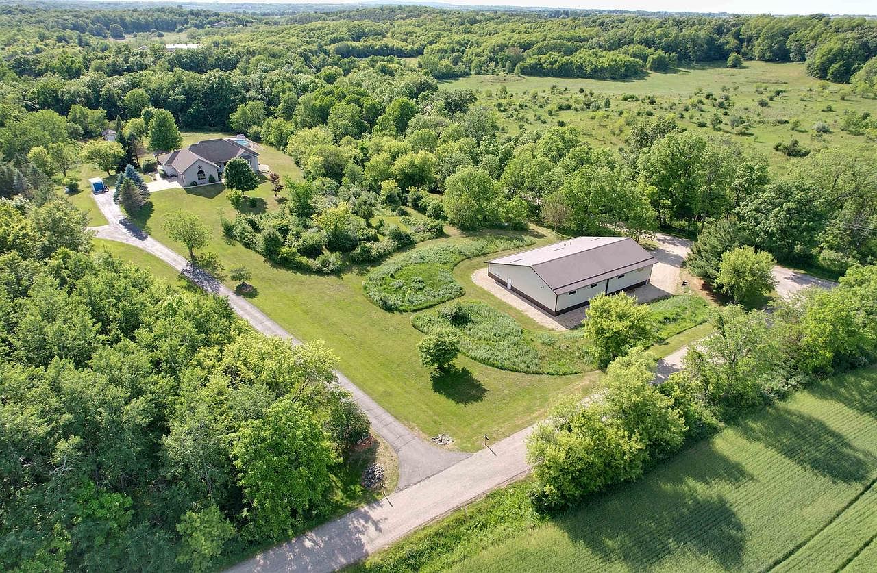 Lower aerial of 1825 Bringold Drive showing the house on the left and the long working shop building on the right, connected by a curving paved driveway across mowed lawn ringed by trees