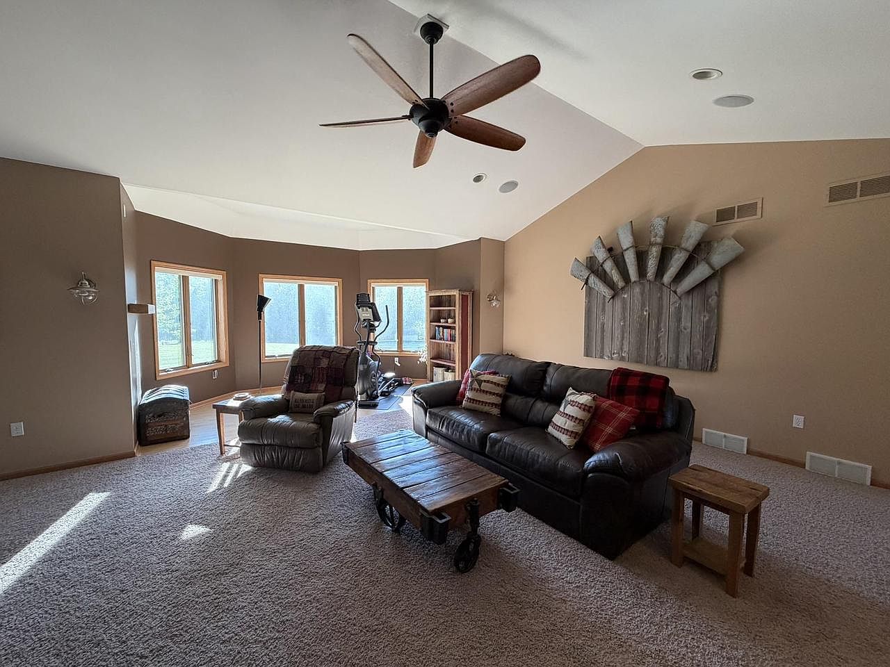 Living room at 1825 Bringold Drive with a soft vaulted ceiling, leather sofa and recliner, a wood plank coffee table on iron casters, a bay of windows on the left, and a salvaged metal wall sculpture above the sofa