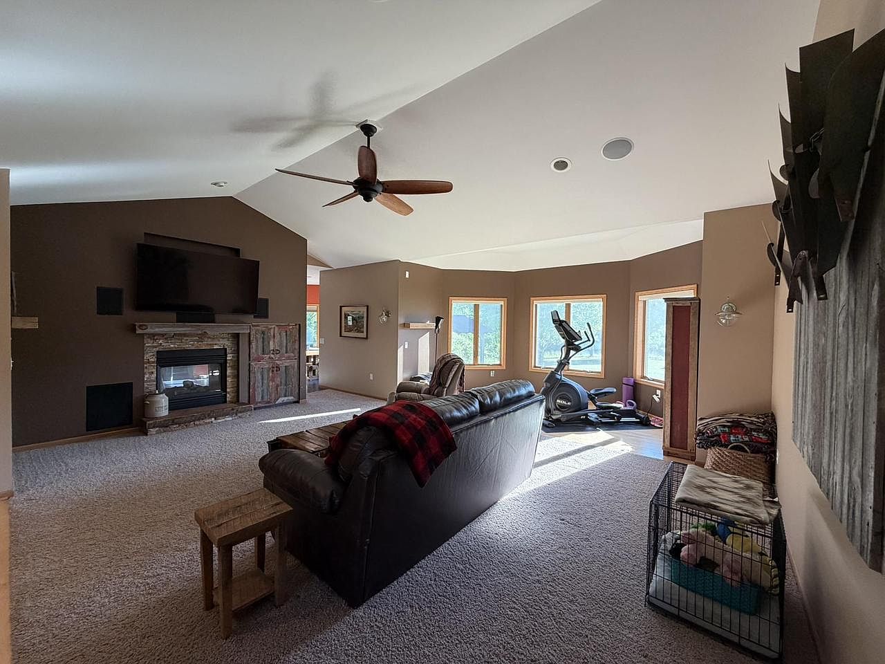 Wide view of the great room at 1825 Bringold Drive with a brown leather sofa center-foreground, the two-sided stacked-stone fireplace and barn-door wood paneling anchoring the far left wall beneath a mounted television, a soft vaulted ceiling lifting overhead, and a bank of windows on the right side wall