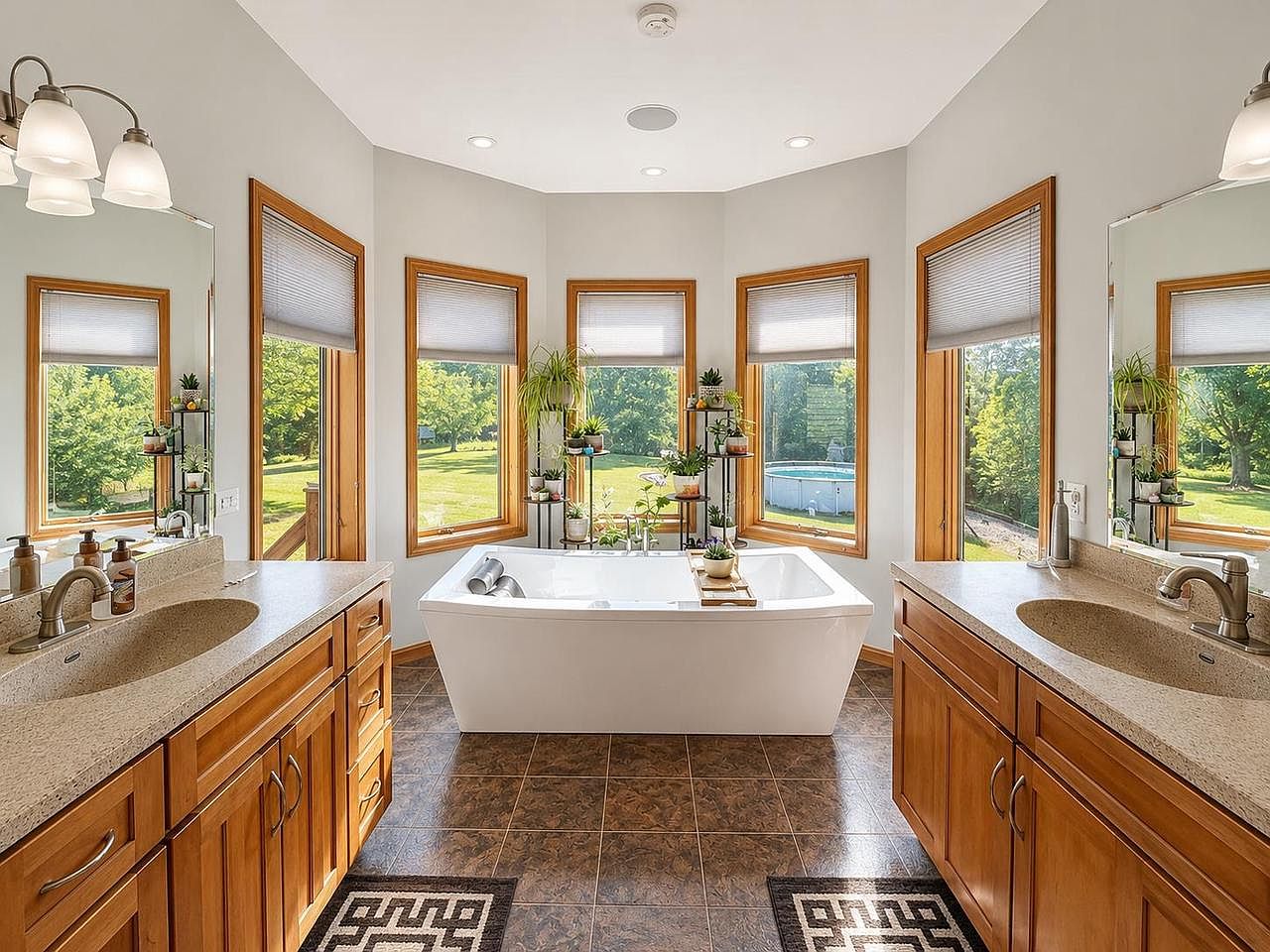 Primary bathroom at 1825 Bringold Drive with a freestanding white soaking tub centered in a bay of five windows that look out across the lawn to the above-ground pool, twin vanities flanking the tub, and a wood-frame mirror over each vanity