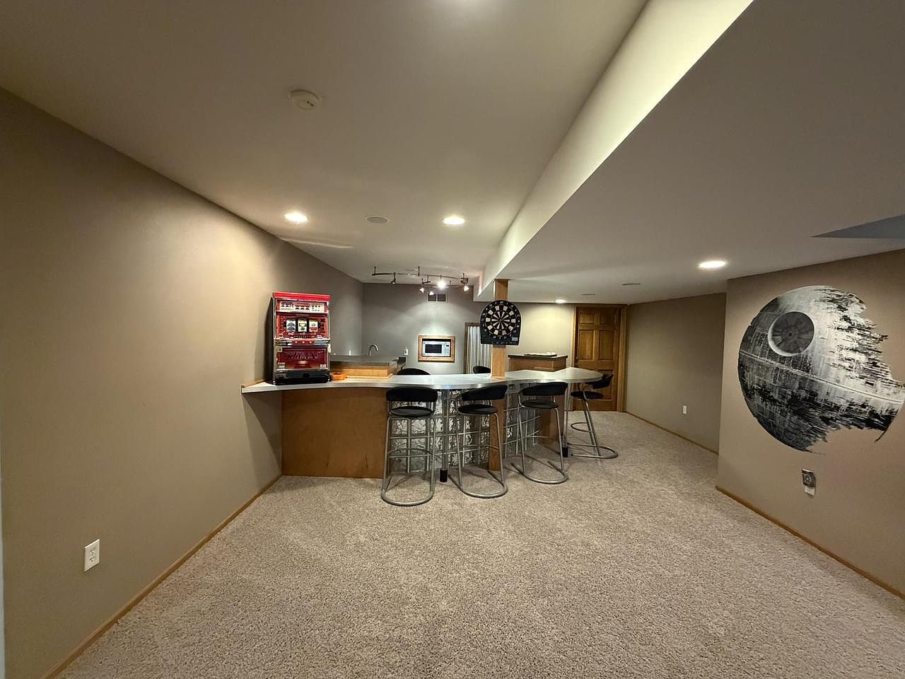 Lower-level bar and rec area at 1825 Bringold Drive with a curved wood bar, four metal stools, an open carpeted lounge behind, and a large Death Star wall mural on the right-hand wall