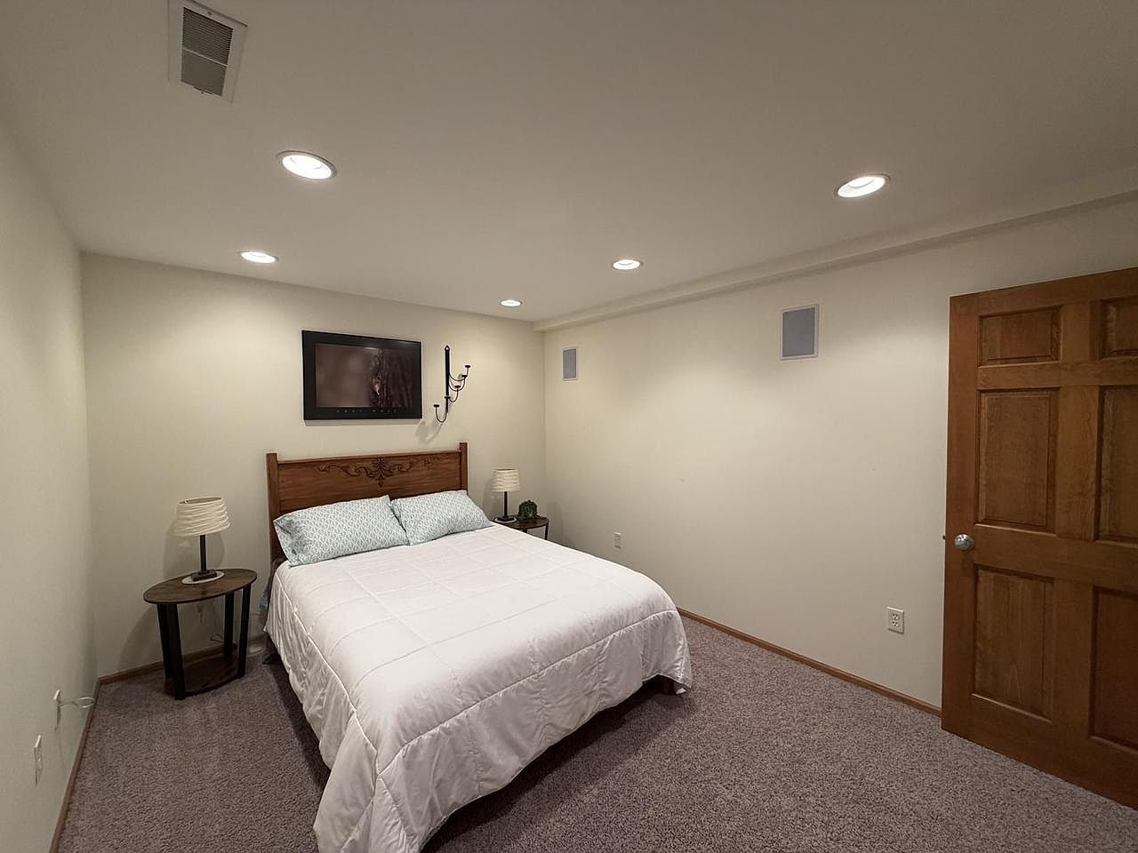 Lower-level guest bedroom at 1825 Bringold Drive: a queen bed with a carved wood headboard and white duvet, twin lamps on round side tables, a flat-screen TV mounted above the bed, recessed ceiling lights, carpet floor, and a six-panel wood door on the right