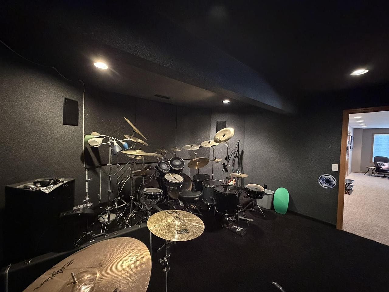 Acoustically-treated lower-level room at 1825 Bringold Drive set up as a drum studio: a full drum kit with cymbals at the center, dark grey wall paneling, recessed lighting, and a small amp at the left