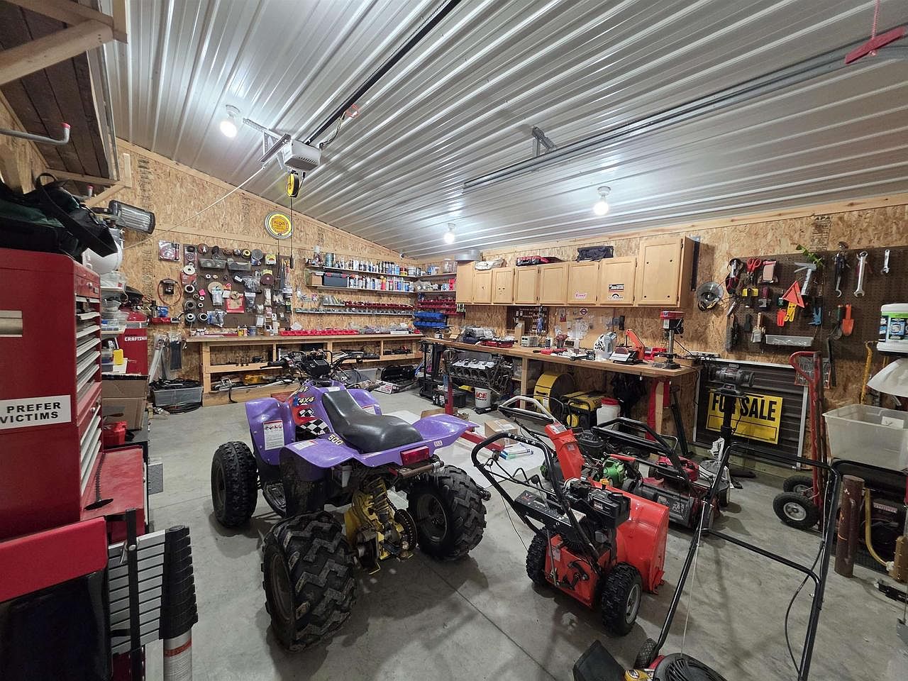 Inside the working shop at 1825 Bringold Drive: a long workbench along the back wall, a purple ATV parked in the center of the concrete floor, a red rolling tool chest at left, a red snowblower at right, and a ribbed metal ceiling overhead