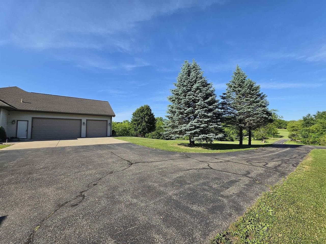 The attached three-car garage at 1825 Bringold Drive seen from the driveway: a hipped-roof home wing on the left with three garage doors, paved driveway curving across the foreground, two tall blue spruce trees on the lawn at right, and an open Wisconsin sky overhead
