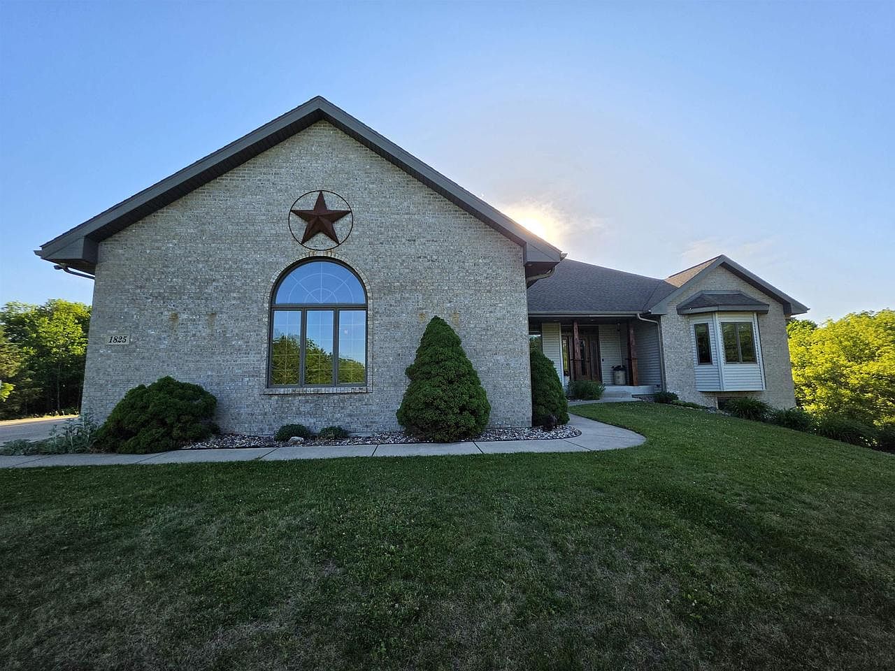 Side elevation of 1825 Bringold Drive at golden hour: a cream brick ranch with a cathedral arched window on the gable end at left, a small metal star above the window, the sun setting behind the gable, in Verona, Wisconsin